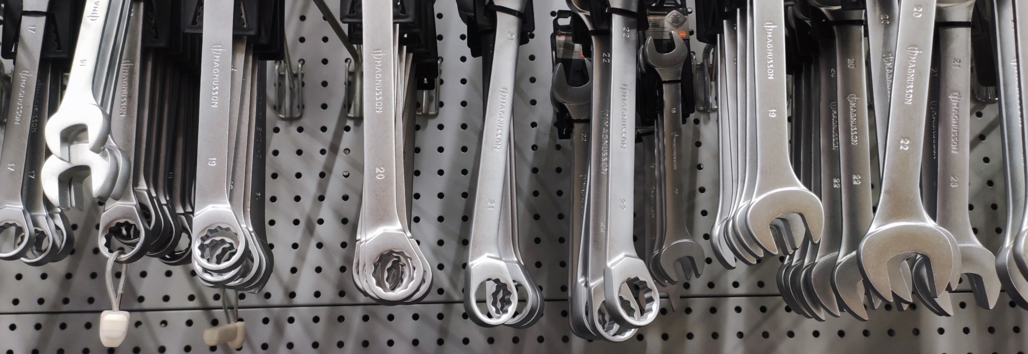Une rangée de clés plates et à cliquet en métal, de tailles variées, suspendues sur un panneau perforé dans un magasin ou un atelier. Chaque clé est étiquetée avec des indications de taille, et la marque ‘Magnusson’ est visible gravée sur certaines clés.